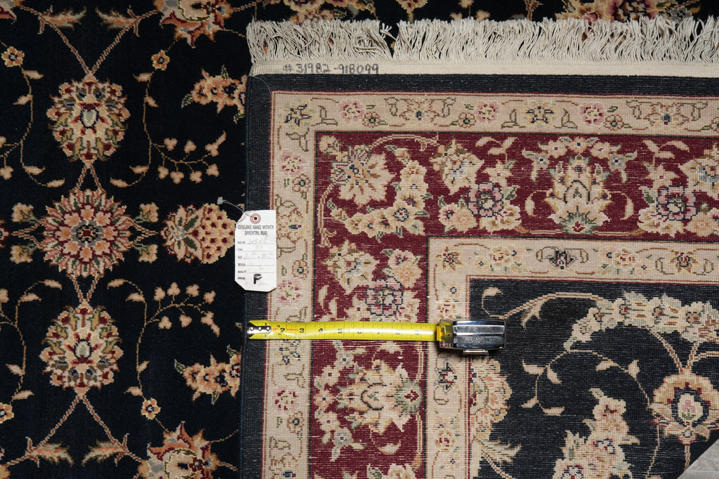 A close-up of a patterned area rug corner with a yellow tape measure laid across it to show dimensions. The rug has a dark navy or black field with beige floral motifs and a burgundy border section.