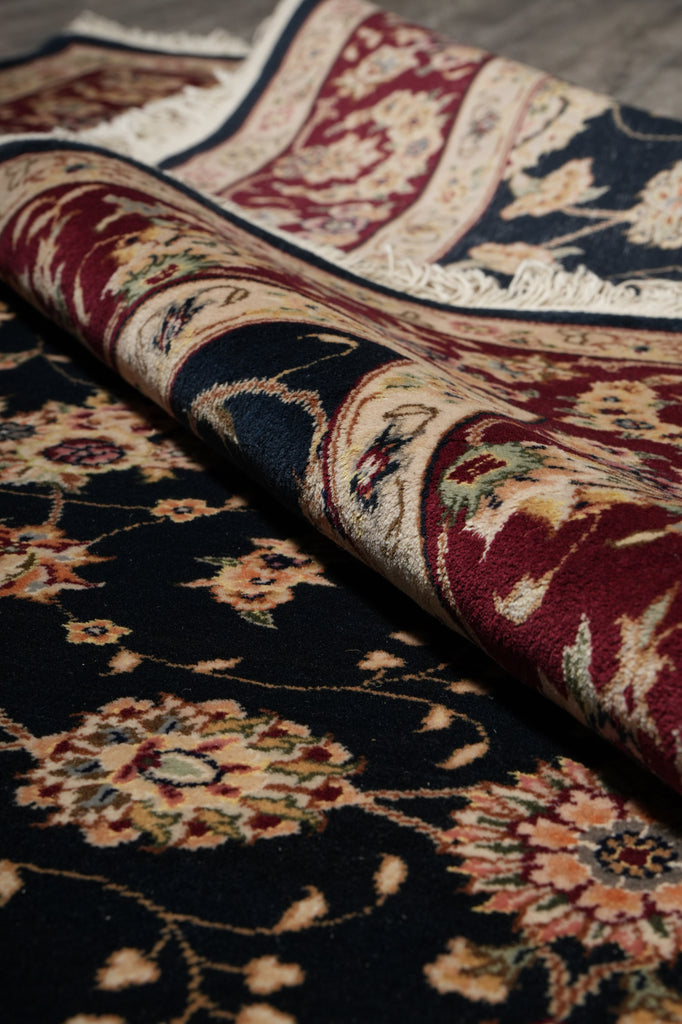 A close-up of a folded traditional area rug showing its detailed floral and vine pattern in deep navy blue, burgundy red, beige, and cream tones.