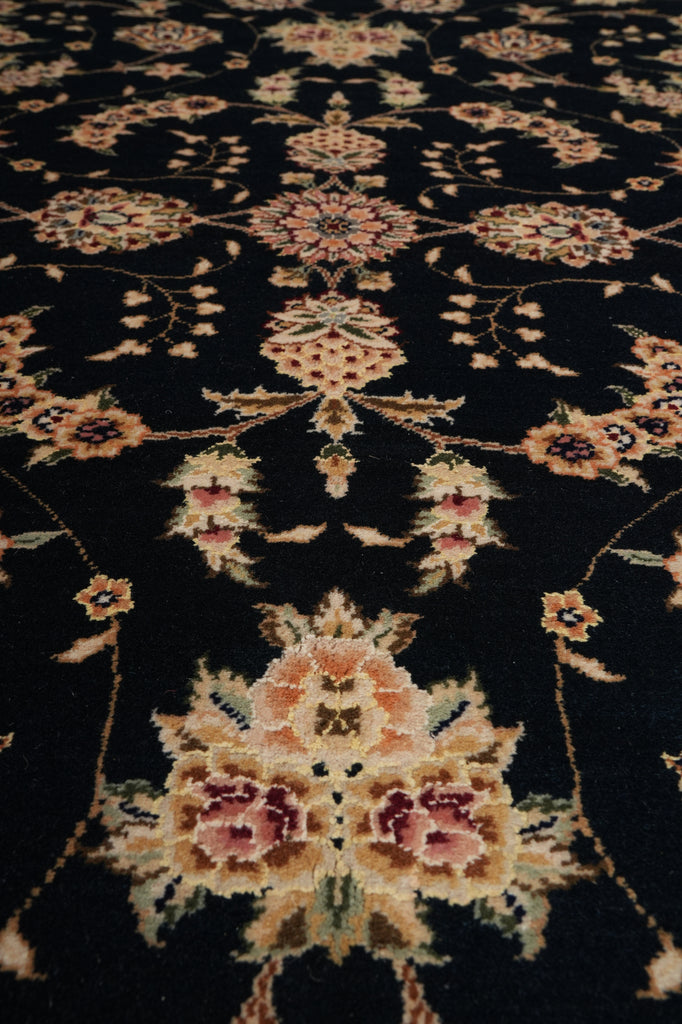 A close-up of a dark black or very deep navy area rug with an ornate traditional floral and vine pattern in beige, muted pink, and olive green.