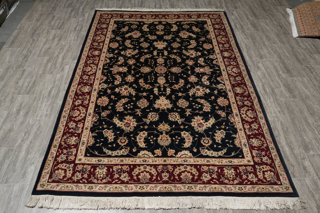 A rectangular traditional area rug with a dark navy blue center, red and beige floral borders, and white fringe, laid out on a gray wood-look floor.
