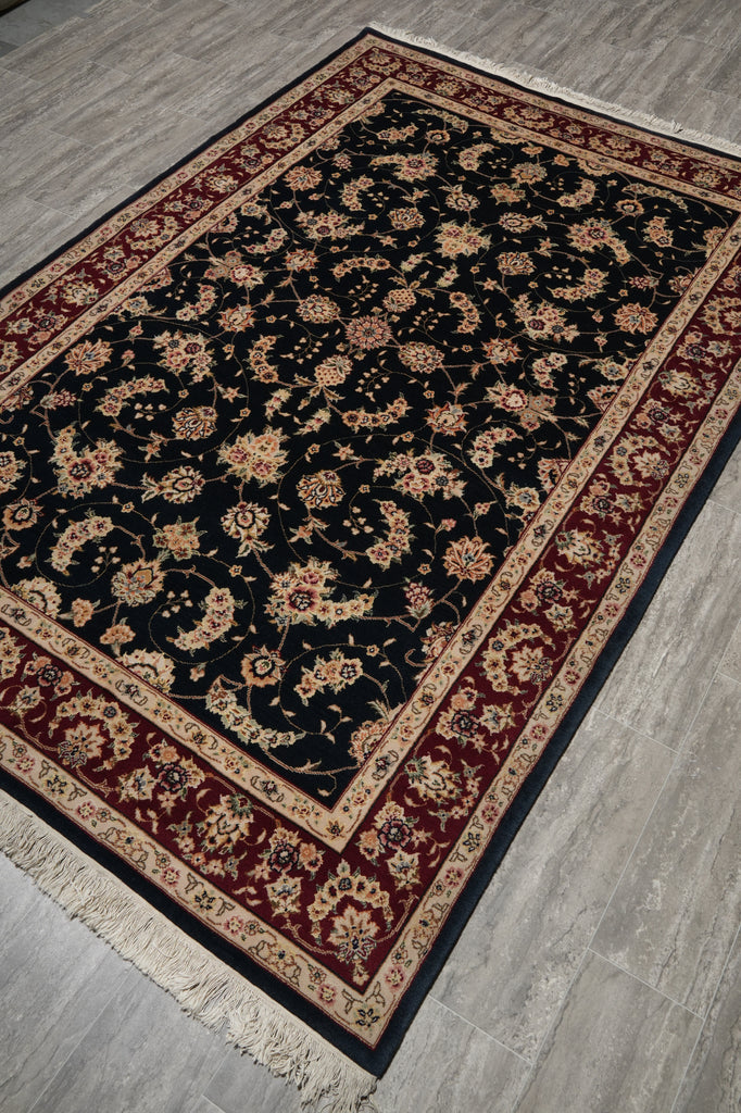 A rectangular area rug with a black central field, ornate floral motifs, and a burgundy and beige border, shown laid out on a gray wood floor.