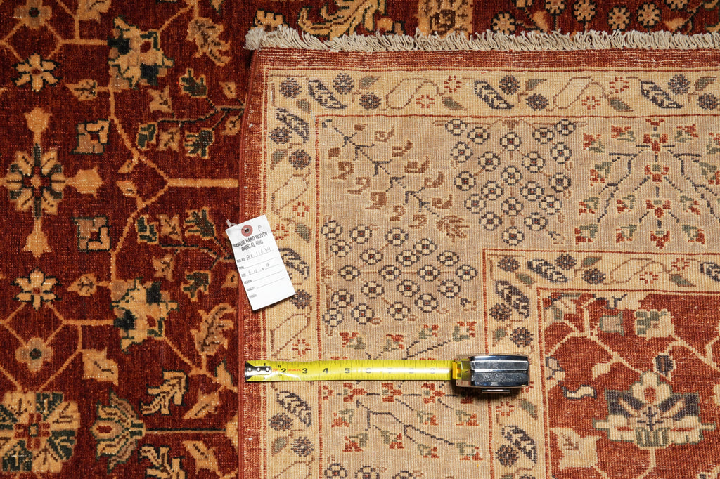 A traditional patterned area rug shown in a top-down view, with a yellow tape measure and a small product tag placed on it for scale. The rug has a warm beige center with red, brown, and muted blue geometric motifs, and a rust-red border.