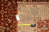 Load image into Gallery viewer, A traditional patterned area rug shown in a top-down view, with a yellow tape measure and a small product tag placed on it for scale. The rug has a warm beige center with red, brown, and muted blue geometric motifs, and a rust-red border.