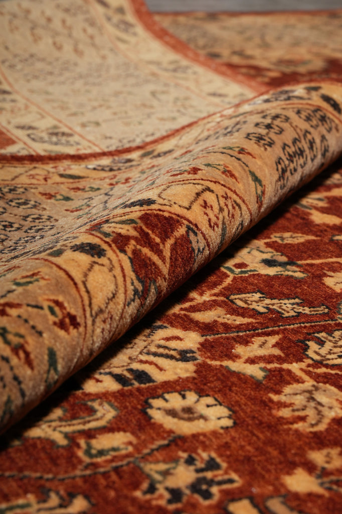 A close-up of a traditional patterned area rug with one end rolled back to show the pile and reverse side, featuring warm rust red, beige, and dark navy floral motifs.