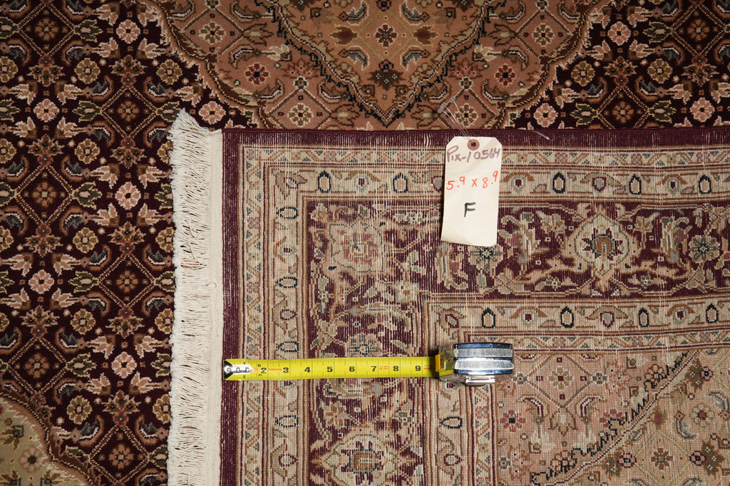 A close-up of an ornate patterned area rug corner with a yellow tape measure and a small handwritten size tag placed on it, showing the rug's pattern and fringe detail.
