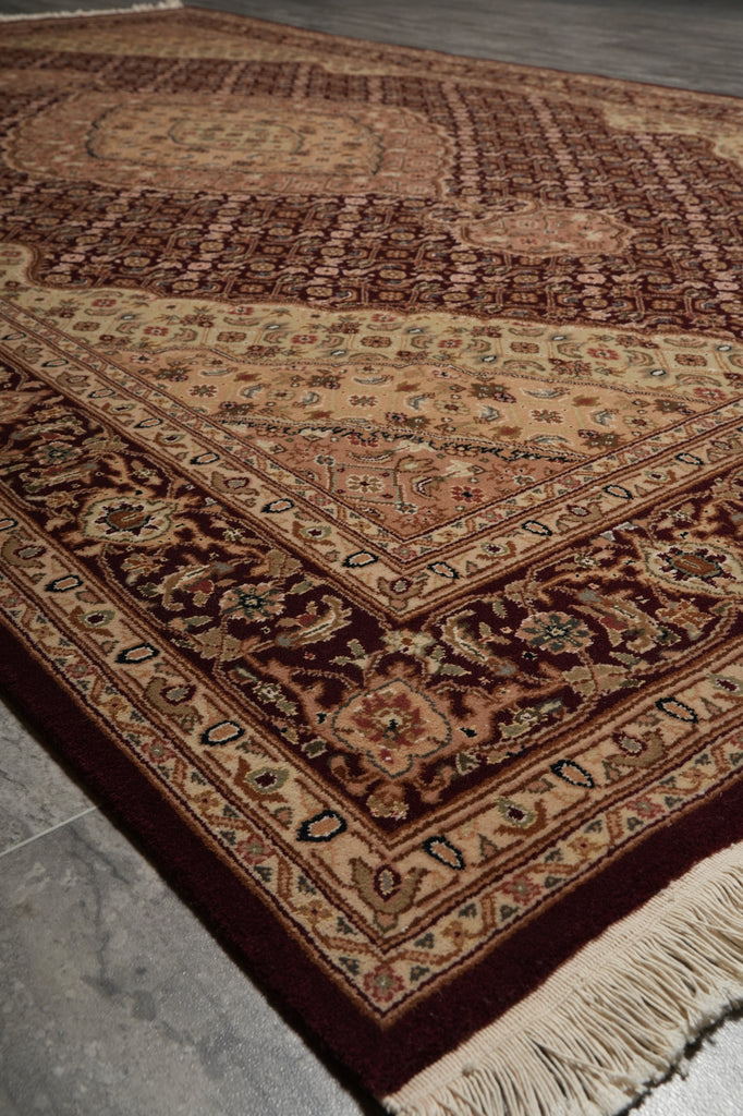 A traditional oriental-style area rug with an intricate floral and geometric pattern, shown laid out on a gray stone floor. The rug features a deep burgundy border, a beige center field, and cream fringe on the corner.