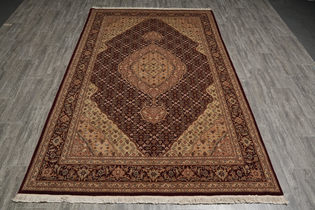 A traditional oriental area rug with an intricate floral pattern, featuring a dark brown central field, beige and gold accents, and a deep red border, laid out on a gray wood-plank floor.