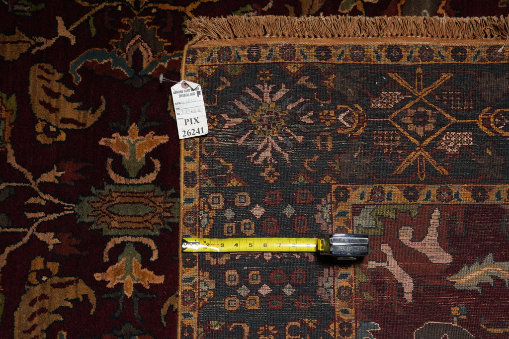 A close-up of a patterned area rug with a dark red field, a dark central panel, and a tan border, shown with a yellow tape measure and a white size tag.