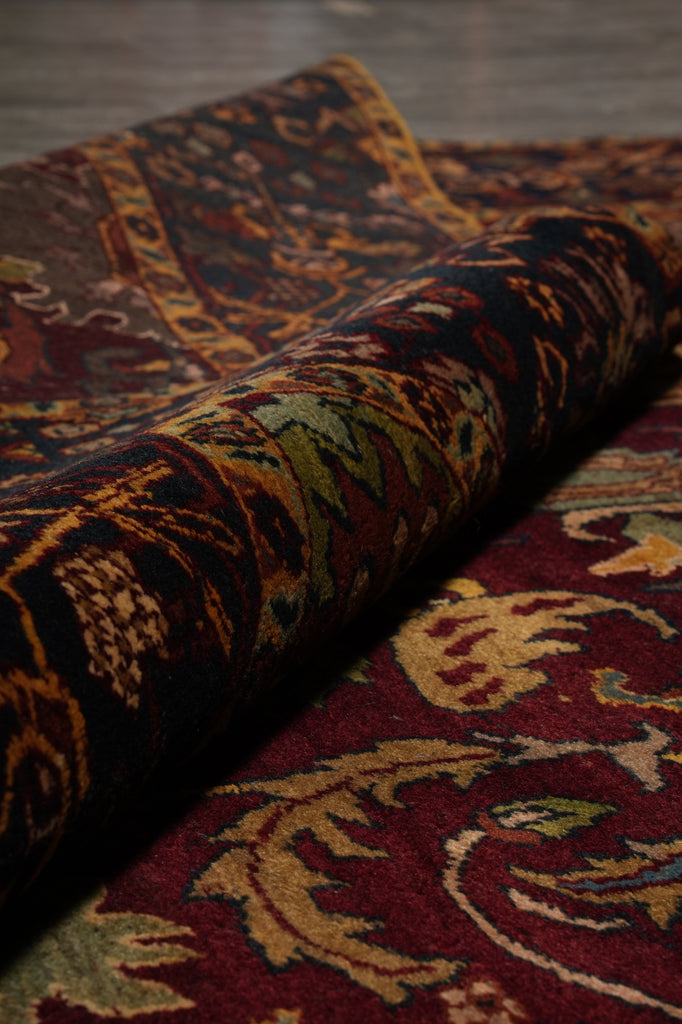A close-up of a rolled area rug showing an intricate traditional pattern in deep red, black, gold, and olive green tones, with the unrolled section displaying a matching floral motif.