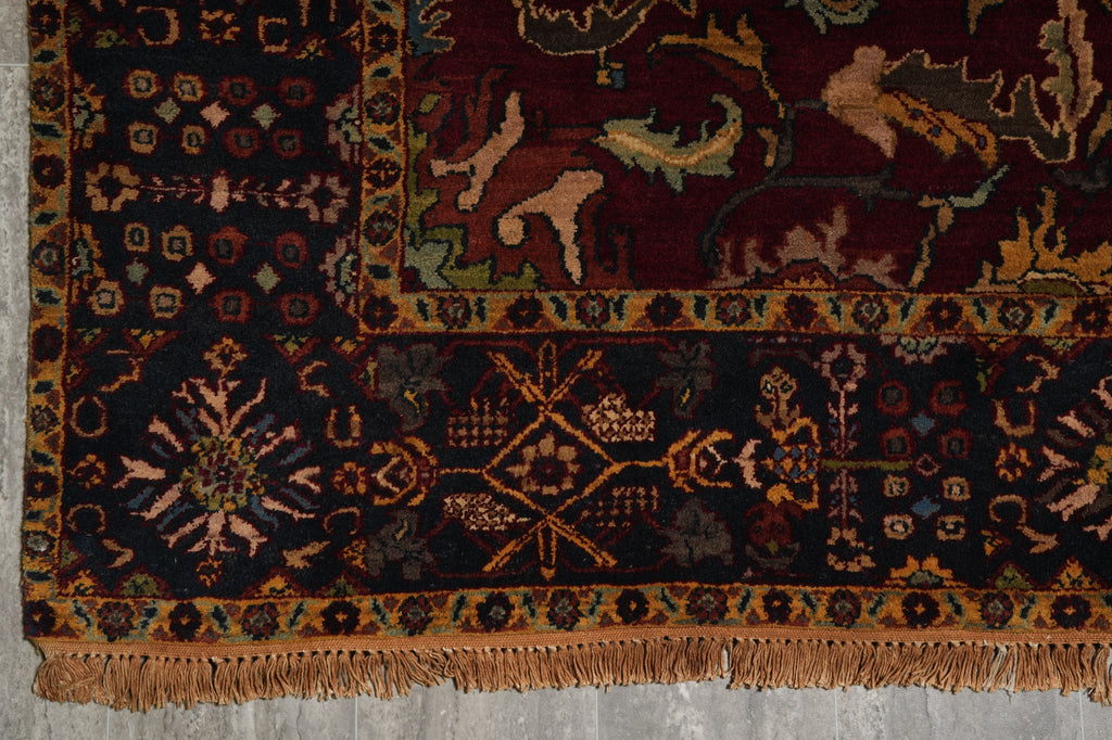 A close-up of an ornate patterned rug corner showing dark navy and burgundy fields with beige, gold, and muted green motifs, plus a fringed edge on a gray floor.