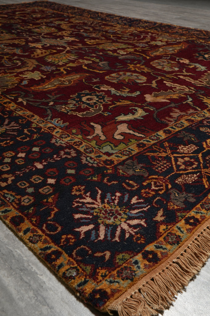 A close-up of a traditional oriental rug with a dark red field, a dark navy blue border, and intricate floral motifs in beige, gold, and green, finished with a fringed edge.