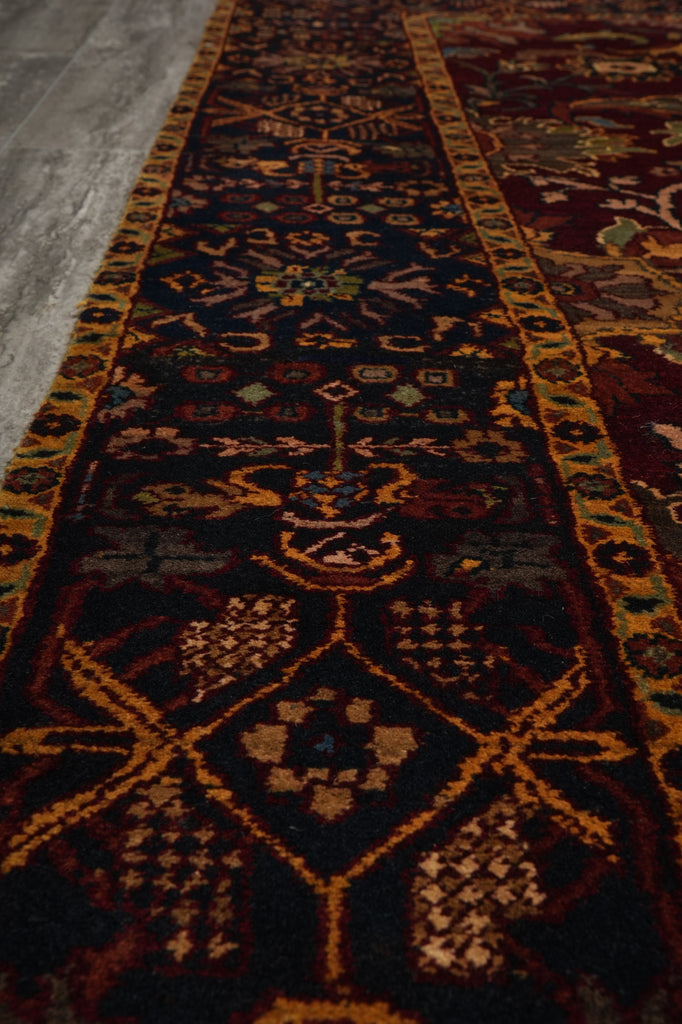 A close-up view of a traditional patterned runner rug with a dark navy blue field, gold and orange borders, and beige geometric motifs, laid out on a gray floor.