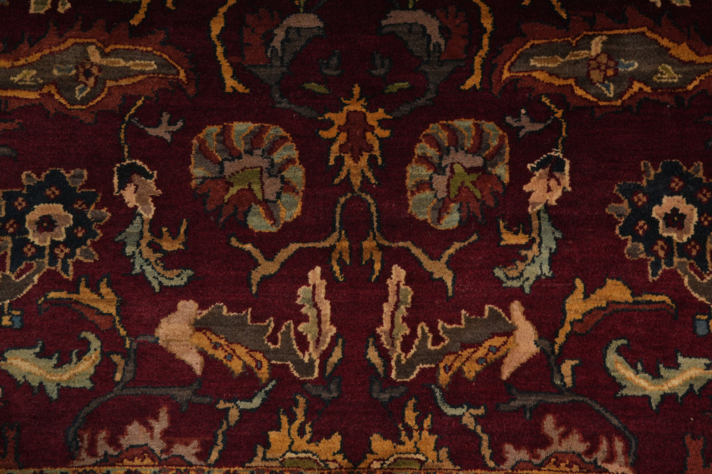 A close-up of an ornate traditional rug with a deep burgundy red field, featuring symmetrical floral and bird motifs in beige, gold, and muted blue-green.