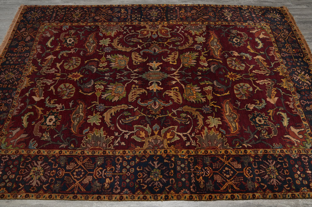 An antique Persian-style area rug with a deep red central field, intricate floral and bird motifs, and dark navy blue borders with gold detailing, laid out on a gray floor.