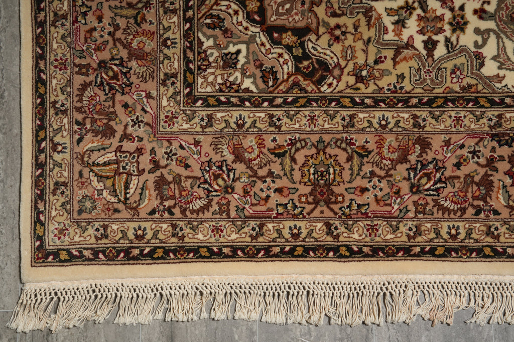 A close-up of an ornate traditional rug corner showing detailed floral and scroll motifs in beige, brown, and muted red tones, with cream fringe along the bottom edge.