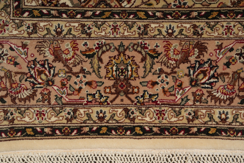 Close-up of an ornate patterned rug showing a central floral medallion and border motifs in beige, brown, red, black, and cream tones, with a cream fringe visible along the bottom edge.