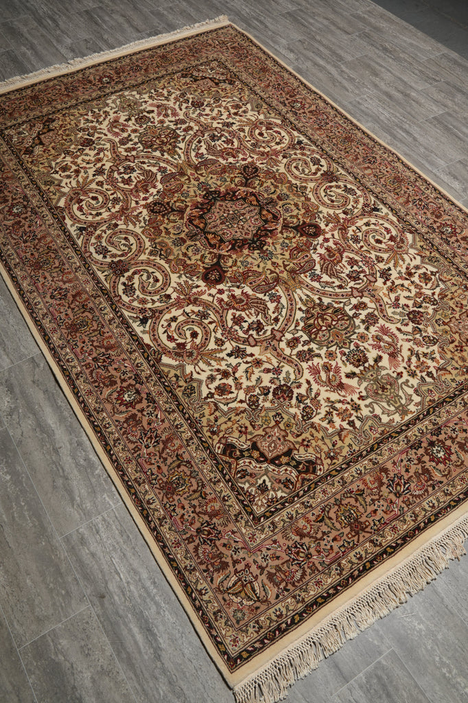 An ornate rectangular area rug with a traditional medallion and floral pattern, shown from an elevated angle on a gray wood-look floor. The rug features a cream center, a warm brown border, and beige fringe.