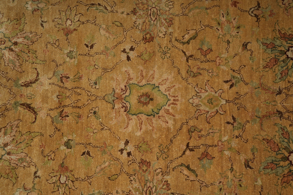 A close-up of an ornate vintage-style rug with a warm tan and ochre background, featuring a central floral medallion and intricate scrolling vine patterns in muted green, brown, and dusty pink.