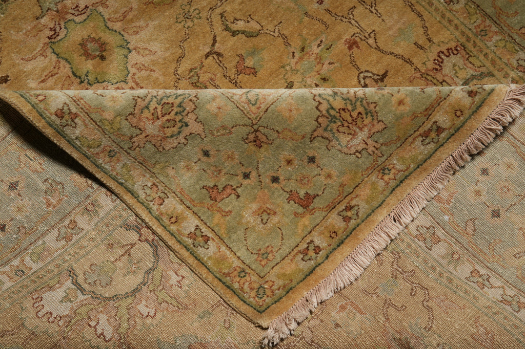 A close-up of an ornate area rug with a folded corner showing its light sage green field, beige border, and cream fringe, over a matching patterned rug.