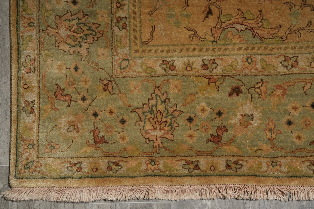 Close-up of an ornate area rug corner showing a muted sage green field with tan, beige, and rust floral motifs, plus a fringed edge.