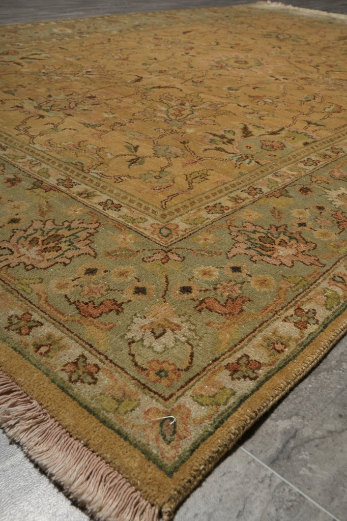 A traditional oriental area rug with a warm mustard yellow field, a sage green inner border, and beige fringe, laid out on a gray tiled floor.