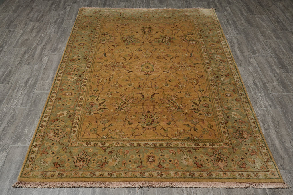 A rectangular area rug with a traditional floral pattern, featuring a warm ochre center and a muted sage green border, laid out on a gray wood-look floor.