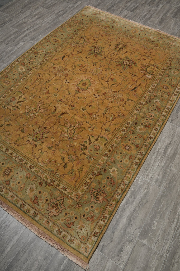 A rectangular area rug with an intricate floral pattern, shown laid out on a gray wood-look floor. The rug features a dominant ochre-yellow center field, a muted sage-green border, and small red and blue accents in the detailed floral motifs.