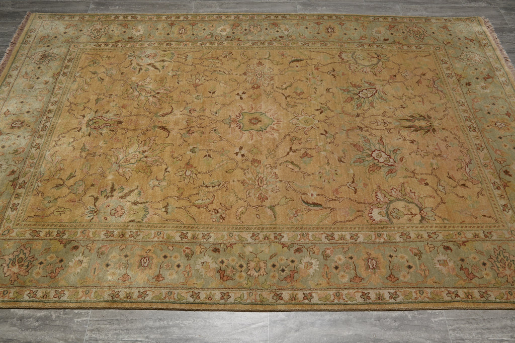 An antique-style oriental area rug with a faded mustard yellow center, a muted green border, and intricate floral motifs in beige, brown, and rust tones, laid out on a gray concrete floor.