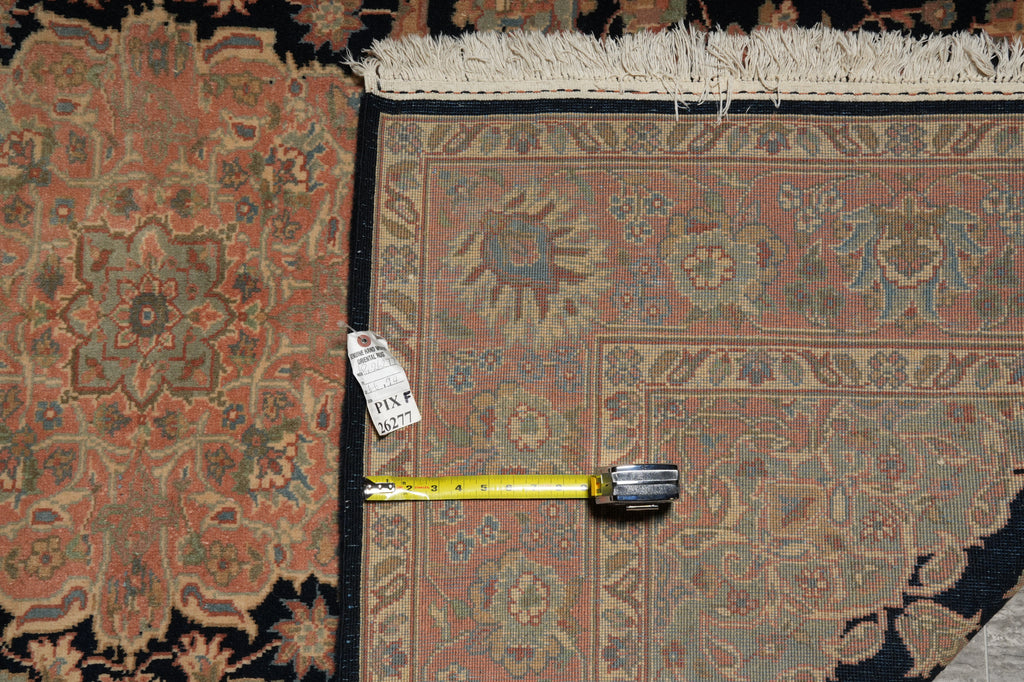 A close-up of a patterned area rug with a corner visible, showing a cream fringe edge, a black border, and a yellow tape measure placed along the inner corner for scale. A small white tag is attached near the tape measure.