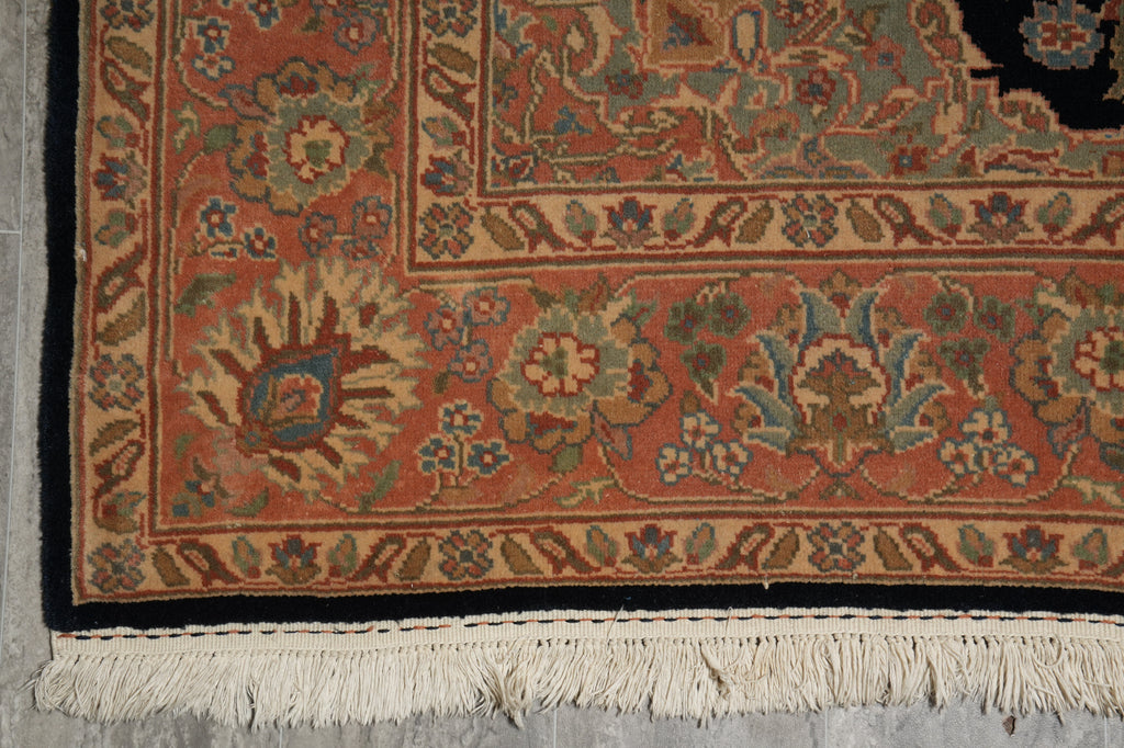 Close-up of an ornate patterned rug corner showing a rust-orange field, cream border, dark navy edge, and white fringe on a gray floor.