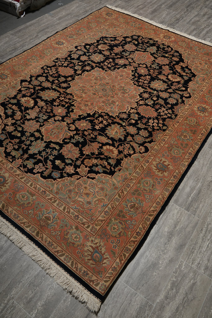 An oriental-style area rug with a complex floral and medallion pattern, shown laid out on a gray wood-look floor. The rug features a black central field surrounded by rust-orange, beige, and muted blue-green details, with a fringed edge.