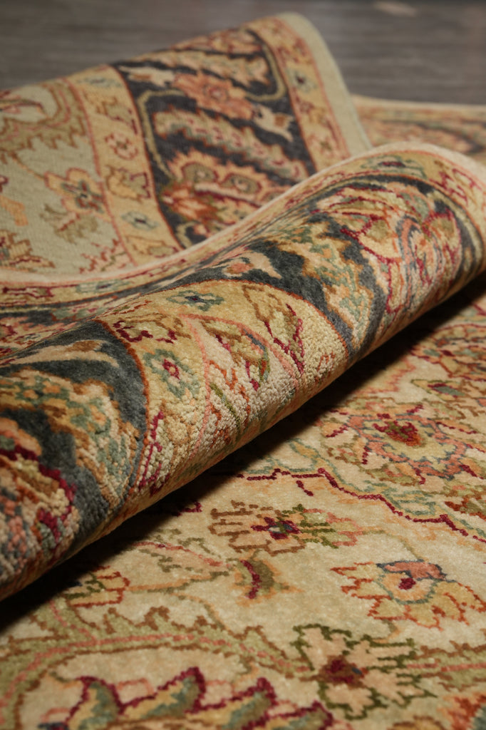 A close-up of a rolled section of a traditional patterned area rug, showing its detailed woven design in beige, green, red, and brown tones over a dark wood floor.