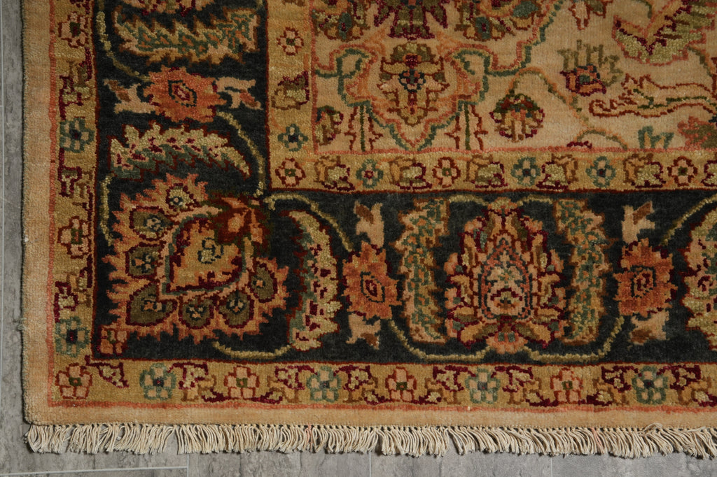 Close-up of an ornate patterned rug corner showing cream fringe, dark navy sections, and warm beige, rust, and green floral motifs.