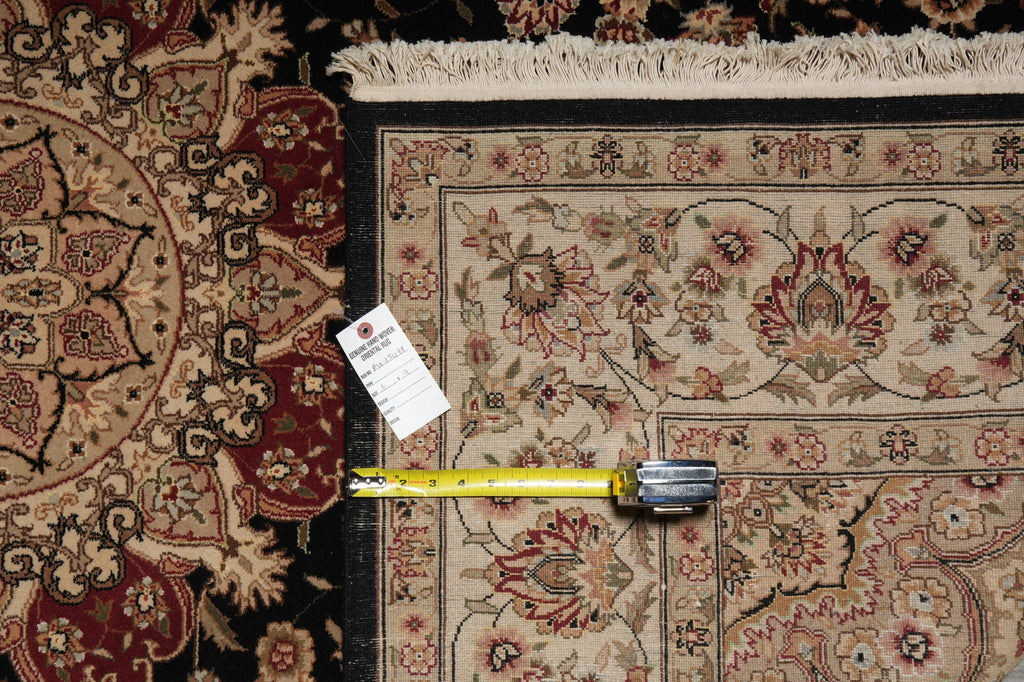 A close-up of a traditional patterned area rug with cream, beige, red, and black tones, shown with a yellow tape measure and a white product tag placed on it for scale.