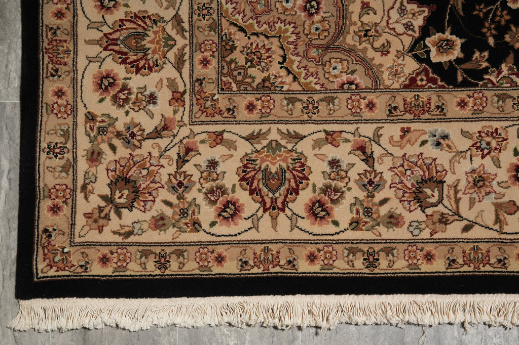 Close-up of an ornate patterned area rug corner showing cream, beige, black, and muted red floral motifs with a fringed edge on a gray floor.
