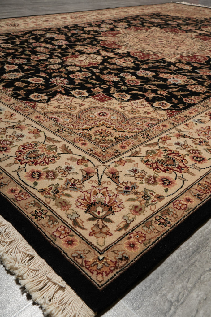 A traditional oriental-style area rug with an intricate floral pattern, featuring a black field, beige borders, and cream fringe, shown laid out on a gray floor.