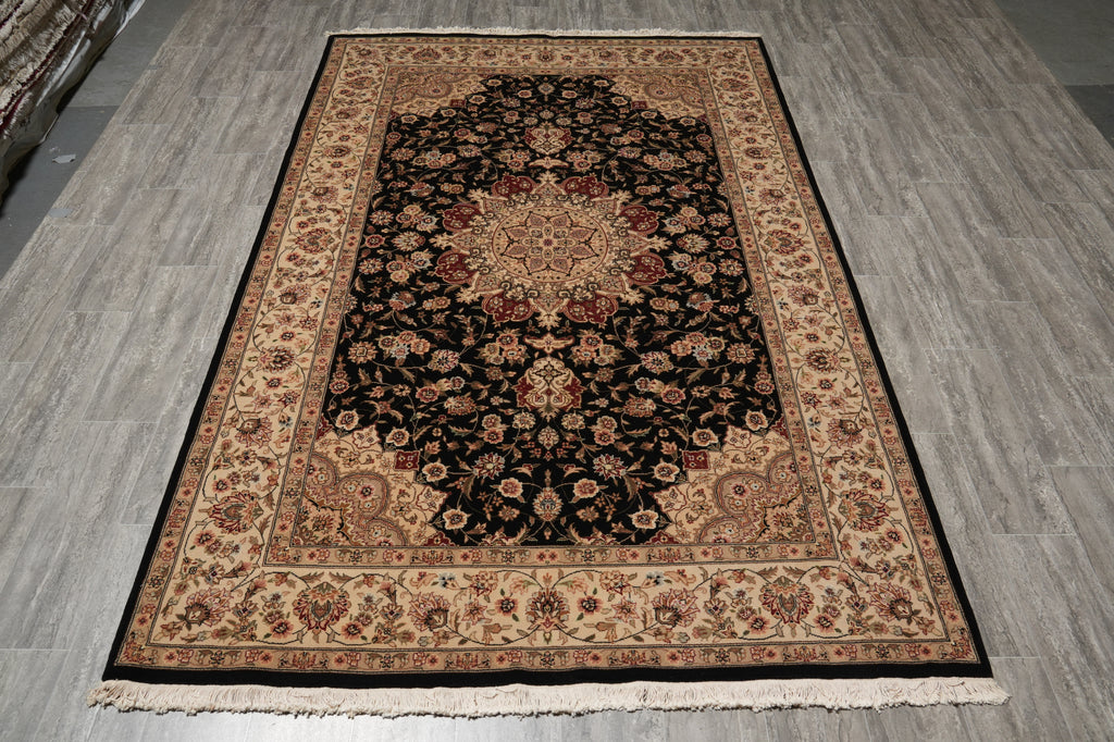 A rectangular area rug with an intricate traditional floral pattern, featuring a black center, beige borders, and red accents, laid out on a gray wood-look floor.