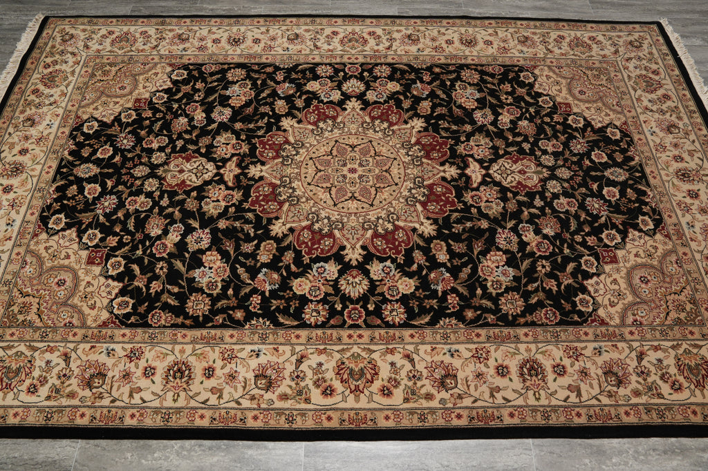 A traditional oriental area rug with an intricate floral pattern, featuring a large black central field, beige borders, and red and gold accents, laid out on a gray tiled floor.