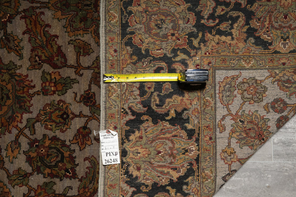 A close-up of a patterned area rug with a vintage-style floral and medallion design in dark navy, rust red, beige, and brown tones. A yellow tape measure is laid horizontally across the rug to indicate scale, and a white inventory tag is attached near the lower left.