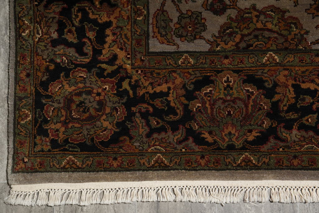 Close-up of an ornate patterned rug corner showing a dark navy and black floral field with green, rust, and beige detailing, plus a white fringe along the bottom edge.