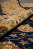 Load image into Gallery viewer, A close-up of a traditional patterned area rug with a rolled section showing the pile and detailed floral motifs in navy blue, beige, and muted green.