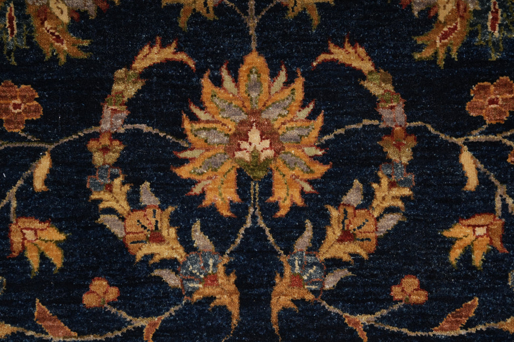 Close-up of a traditional patterned rug featuring a central floral motif in warm gold, orange, and beige tones on a dark navy blue background.
