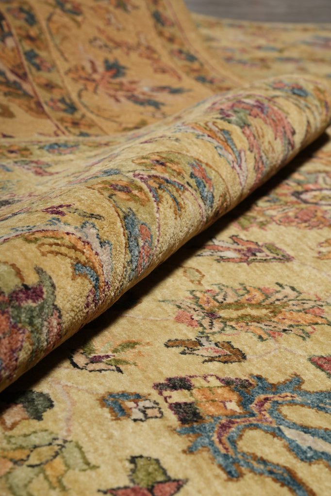 A close-up of a rolled section of a traditional patterned rug showing its detailed floral motifs and pile texture in warm beige, gold, and muted blue, green, and pink tones.