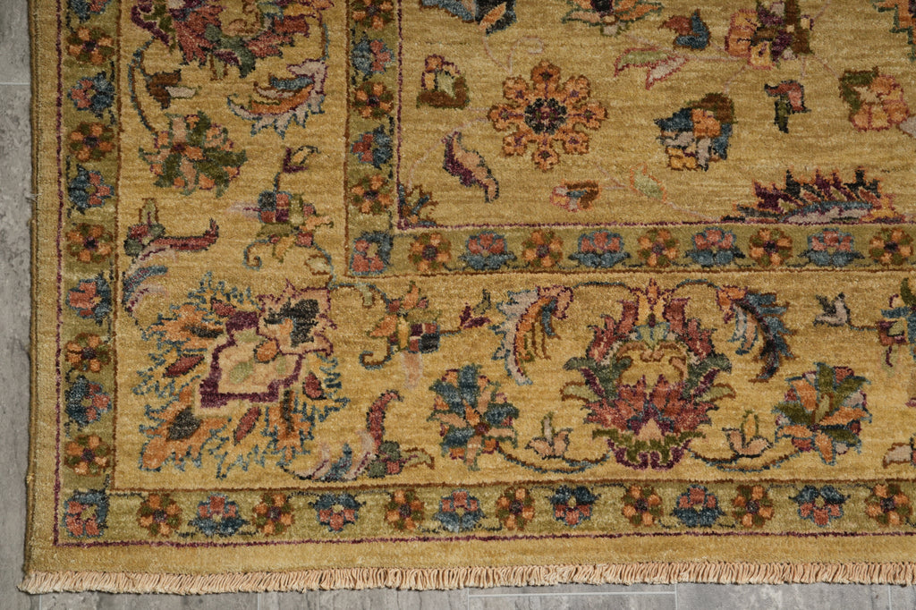A close-up of an ornate traditional area rug with a warm beige and ochre base, featuring intricate floral and botanical motifs in muted red, blue, green, and brown, finished with a fringed edge.