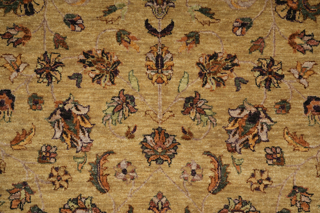 A close-up of a woven rug with a floral and vine pattern on a warm golden-beige background. The design features muted brown, green, and cream motifs throughout.