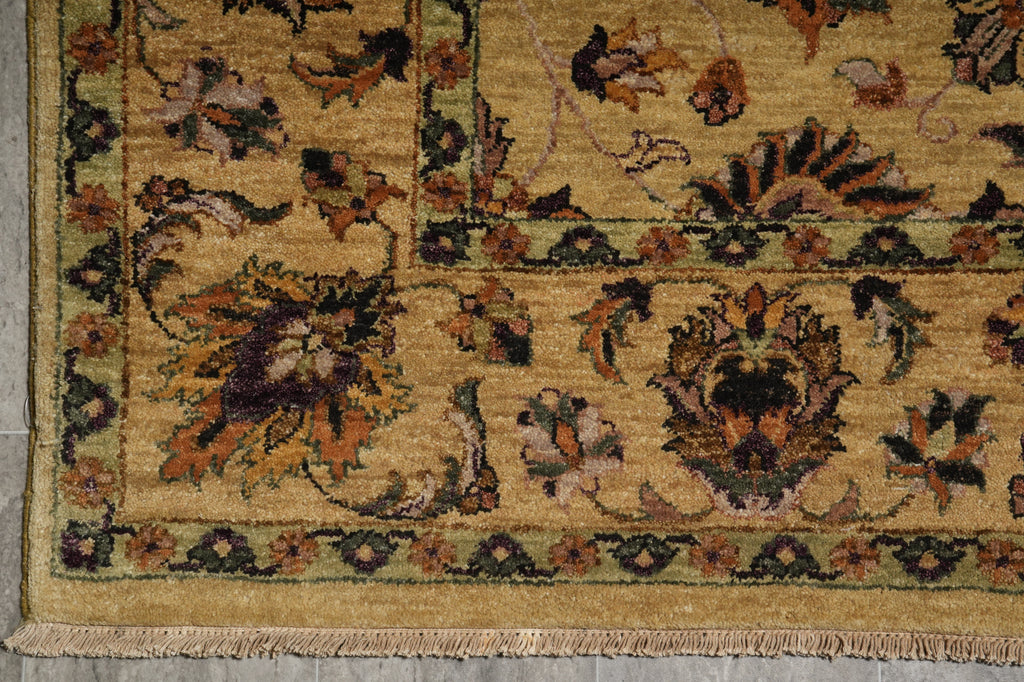 Close-up of a traditional patterned rug corner showing a beige and gold field with floral motifs in brown, green, and rust tones, plus a fringed edge.