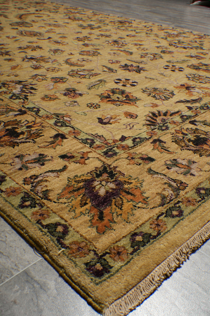 A close-up of a traditional area rug with a warm beige and mustard yellow base, featuring an intricate floral pattern in brown, orange, and green, with a fringed edge on a gray tiled floor.