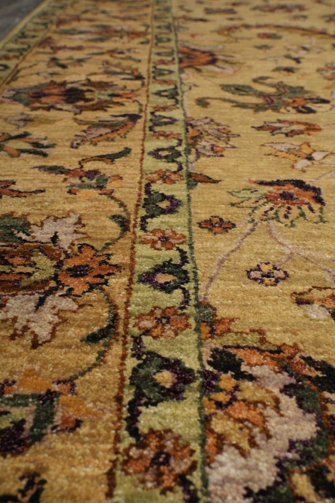 A close-up view of an ornate area rug with a warm golden-yellow base, featuring a central vertical border and floral motifs in green, brown, orange, and cream.