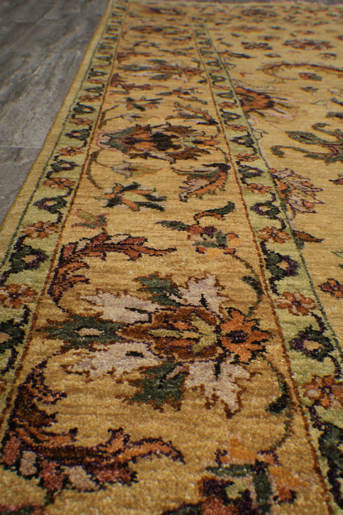 A close-up view of a traditional patterned area rug with a warm mustard yellow base, featuring floral motifs in brown, green, beige, and muted orange, bordered by ornate patterns.