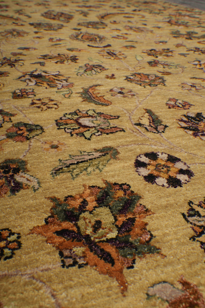 A close-up view of a textured area rug with a warm mustard yellow base and an intricate floral pattern in orange, green, brown, and cream.